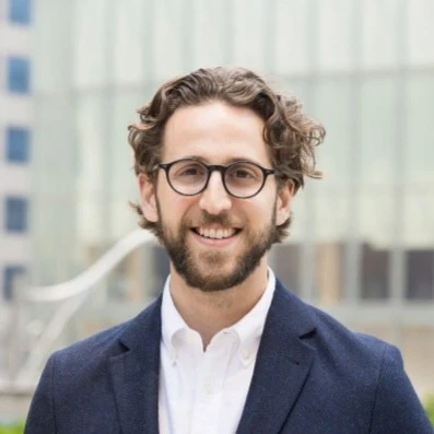 Smiling man with glasses and a beard, wearing a suit jacket, stands outside in front of a modern building.