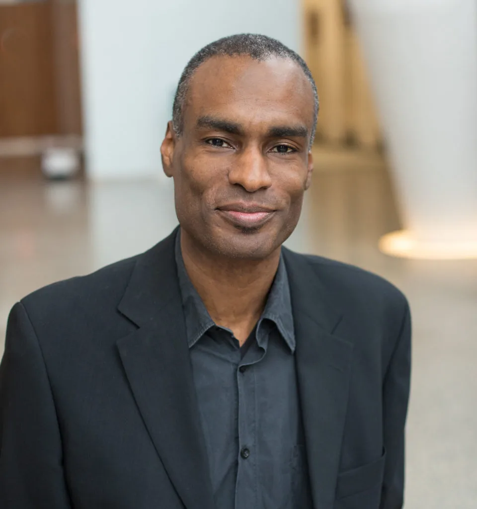 Man in a black suit and shirt smiles slightly, standing indoors with a blurred background.