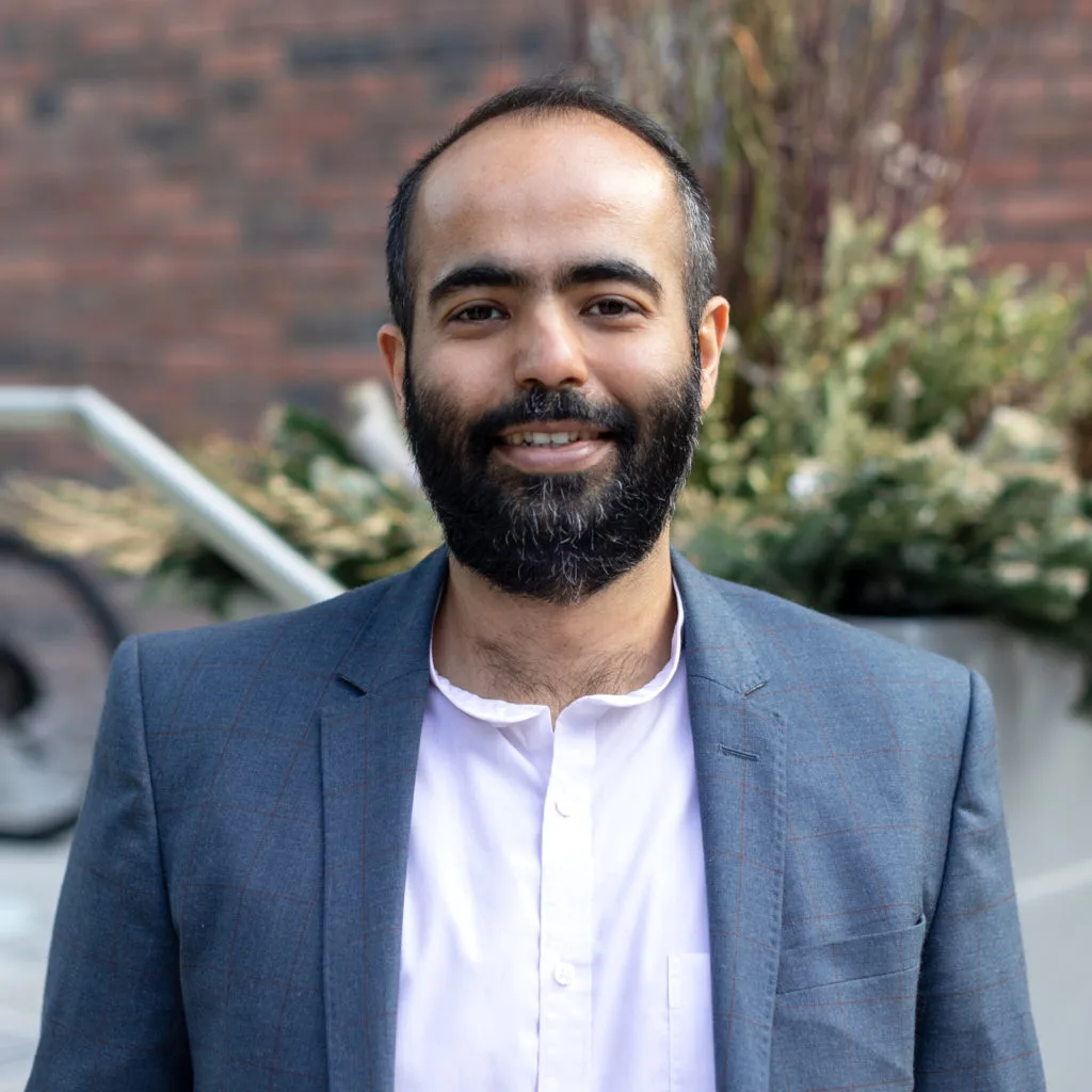 Man with a beard in a blue blazer and white shirt smiles outdoors in front of plants and a brick wall.