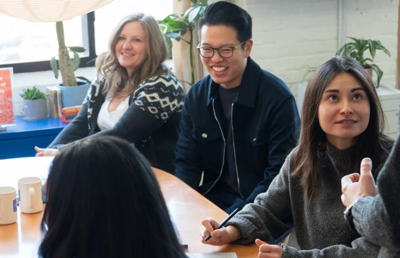 Four people sit at a table in a bright office, smiling and listening during a meeting.