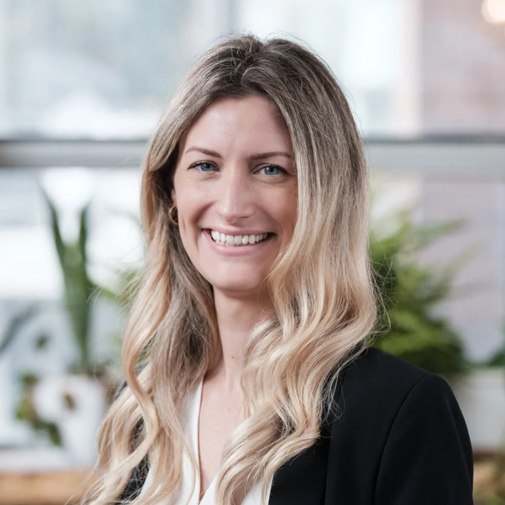Smiling woman with long blonde hair in a black blazer, standing indoors with plants in the background.