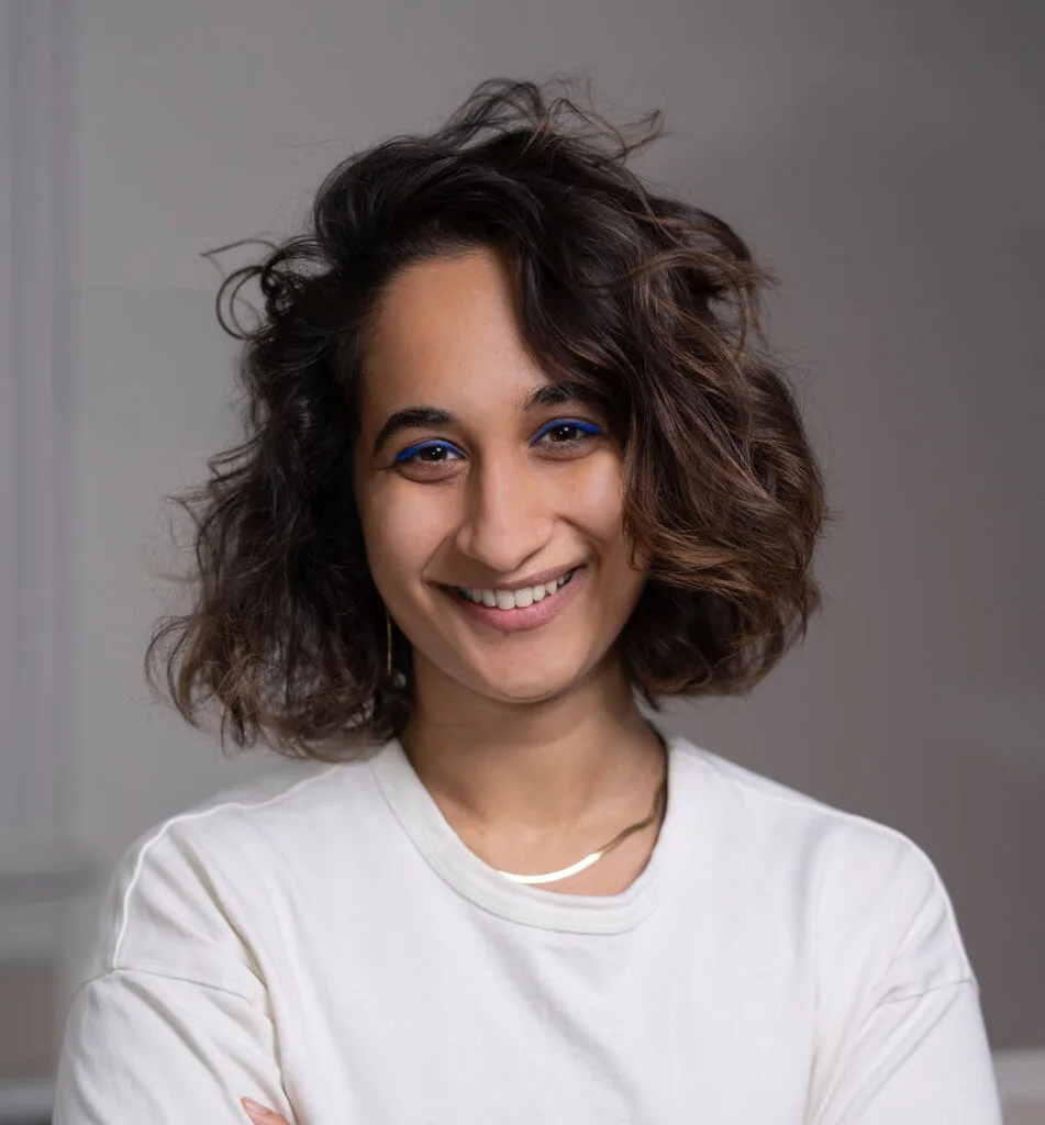 Smiling person with wavy brown hair and blue eyeliner wearing a white shirt, standing in front of a gray background.