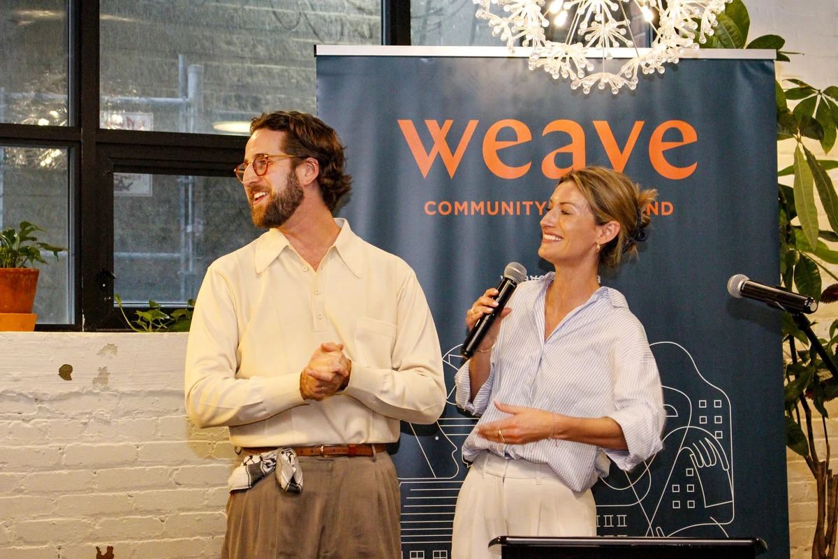 Two people stand smiling with microphones in front of a weave community fund banner indoors.